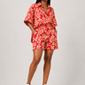 Woman wearing a red and cream Ankara print matching shirt and shorts set with wide kimono sleeves and high waist tailored shorts styled with white sneakers against a grey background