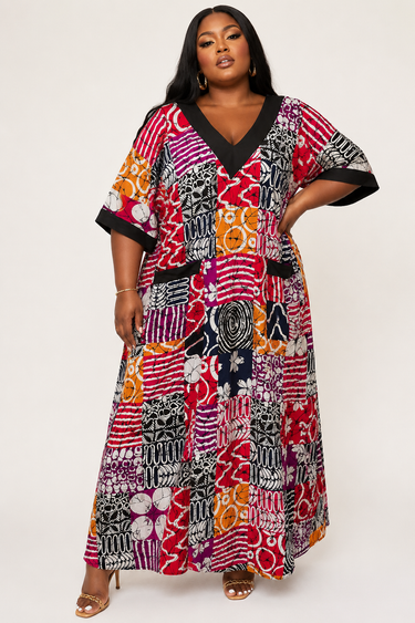 Plus size model wearing a vibrant Ankara patchwork maxi dress with black V neck trim and short kimono sleeves styled with gold heeled sandals against a white background