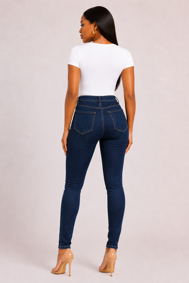 Woman wearing deep indigo dark wash high waist skinny jeans with slim fitted tapered leg golden contrast stitching and clean saturated color styled with a white bodysuit and nude strappy heeled sandals against a light grey background