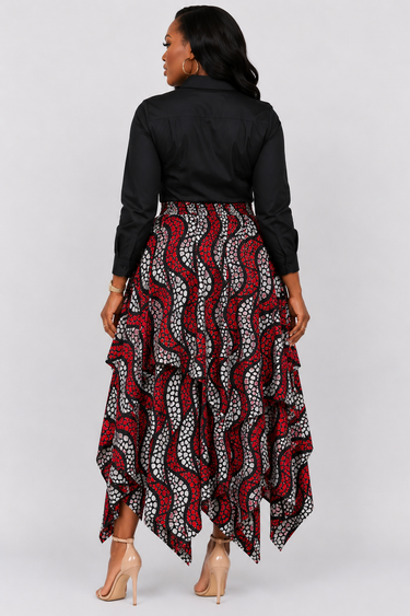 Woman wearing a bold red black and grey Ankara wave print handkerchief maxi skirt with high waist and layered cascading ruffle hem styled with a black button down shirt and nude heeled sandals against a grey background